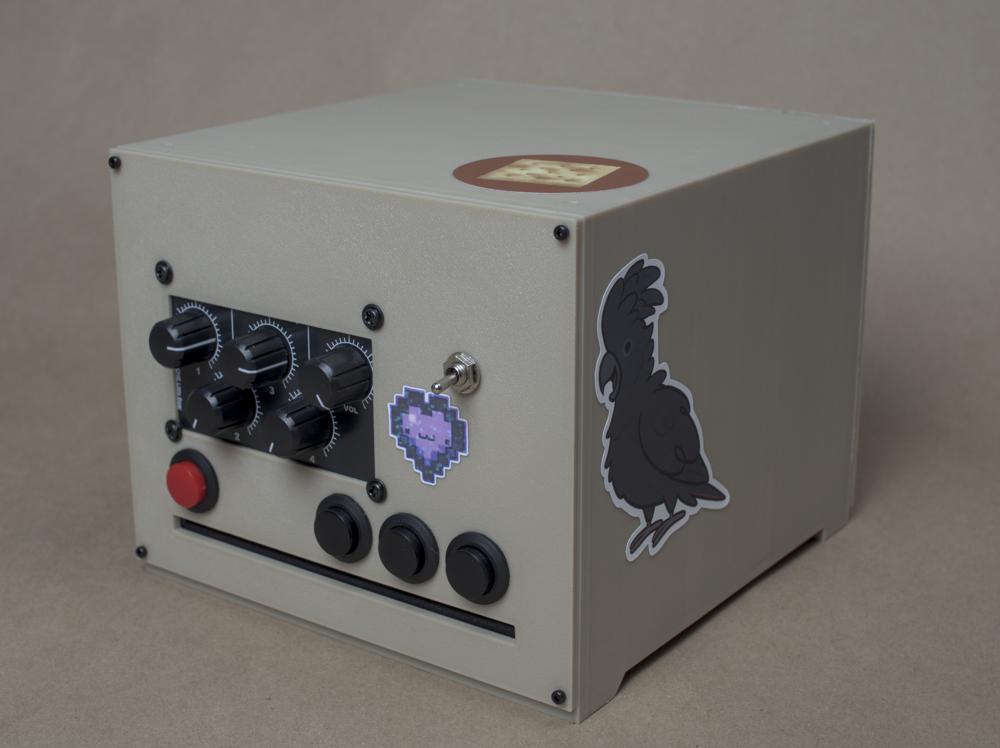 A 3d printed box decorated with stickers. On the front side 4 buttons are present, a cd sized slit, a 5 dial, and a switch